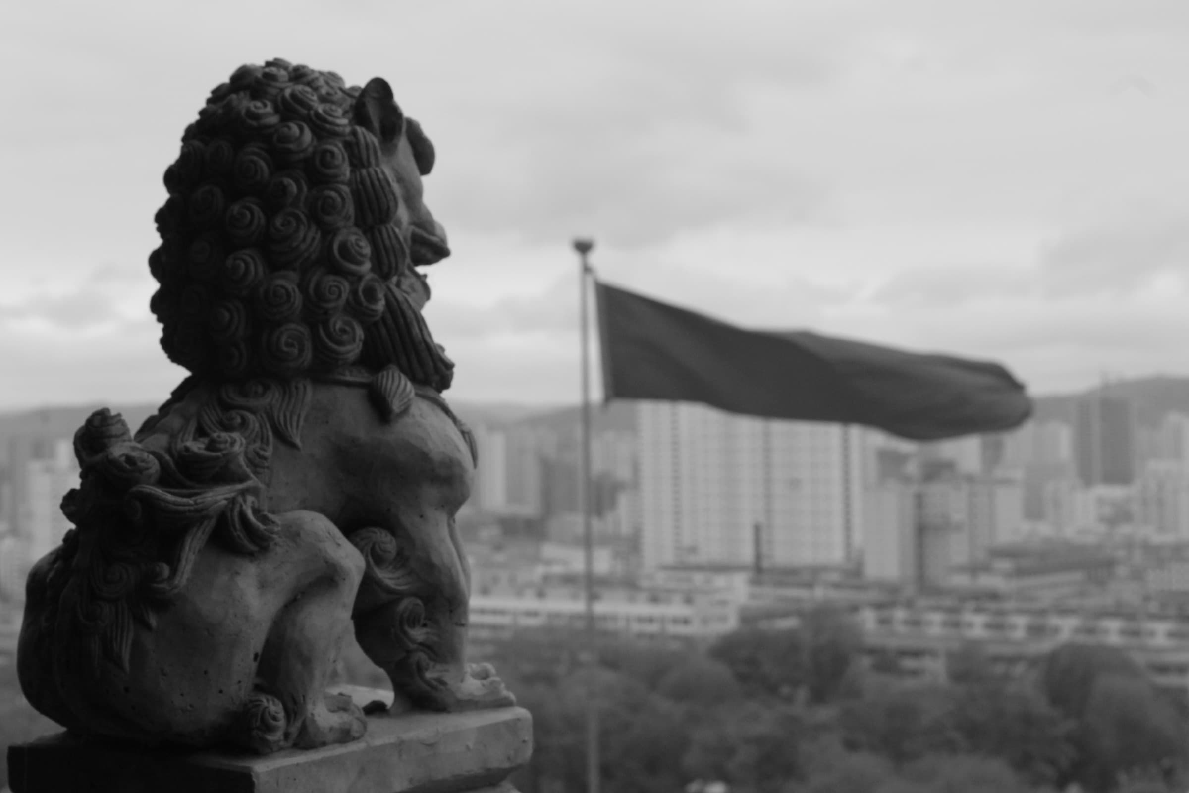 Lion de pierre monumental dressé au-dessus de la ville ; derrière lui, un grand drapeau claque au vent au-dessus d’un océan d’immeubles.