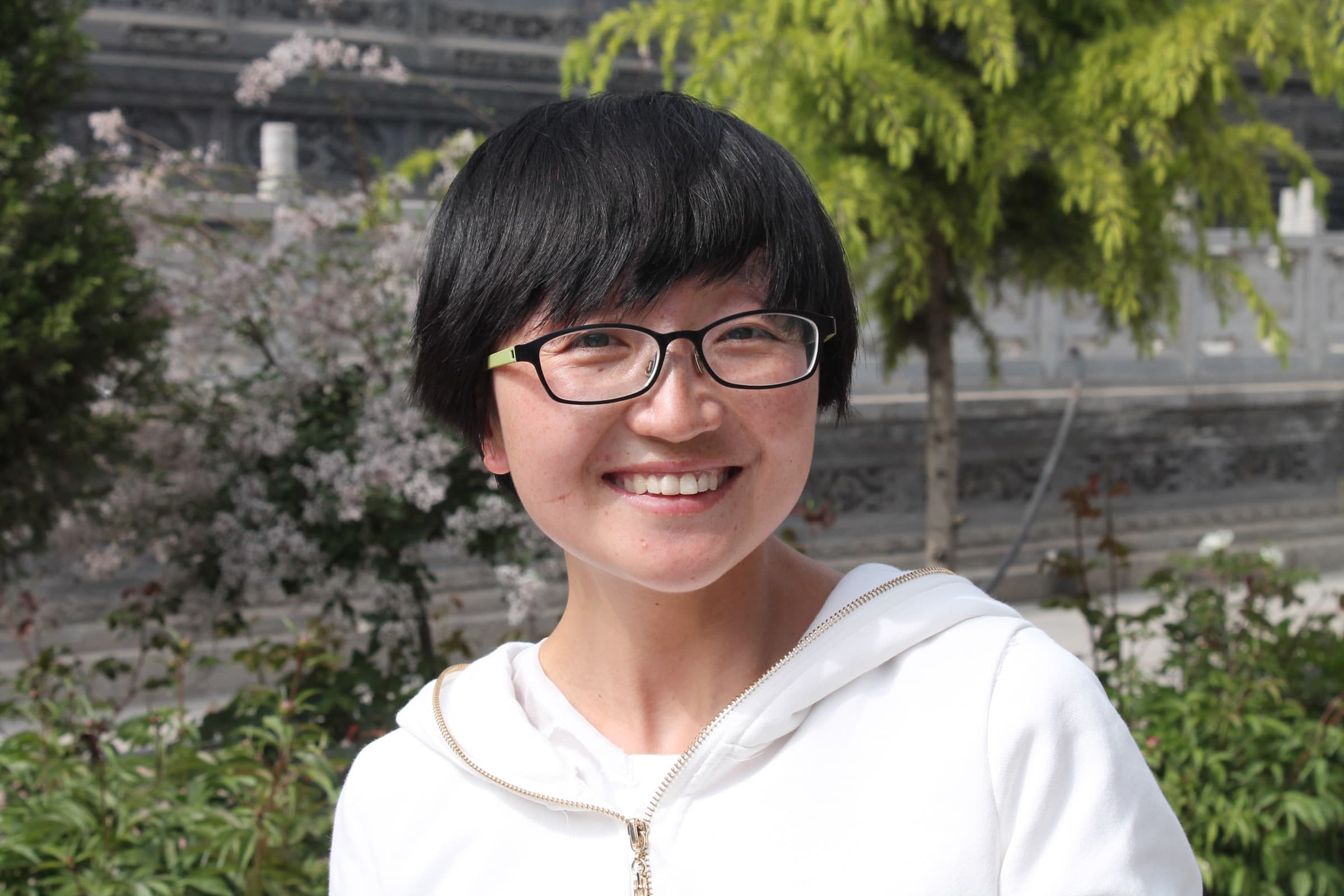 Jeune femme aux cheveux courts et lunettes, en sweat blanc à capuche, souriant largement devant un arbre en fleurs.