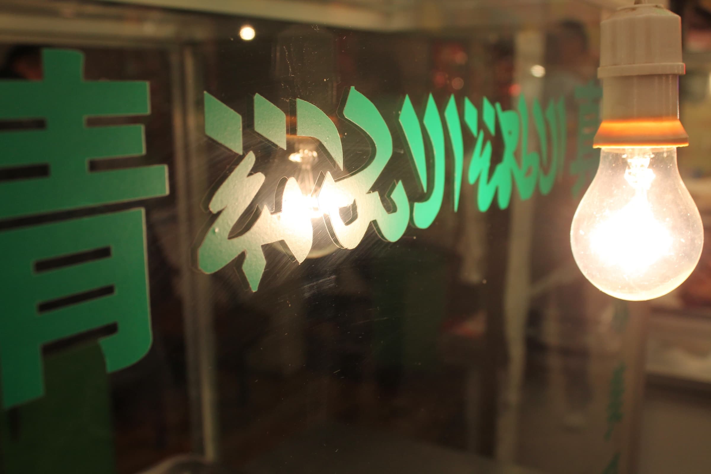 Enseigne lumineuse verte où un caractère chinois voisine avec une calligraphie arabe, éclairée par une ampoule nue.