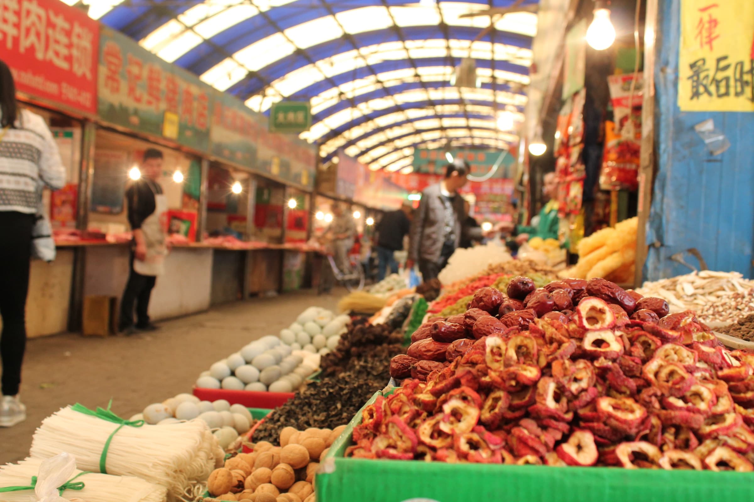 Longue allée de marché couvert : bacs débordants de piments séchés, de fruits confits et de racines, sous une verrière en berceau.