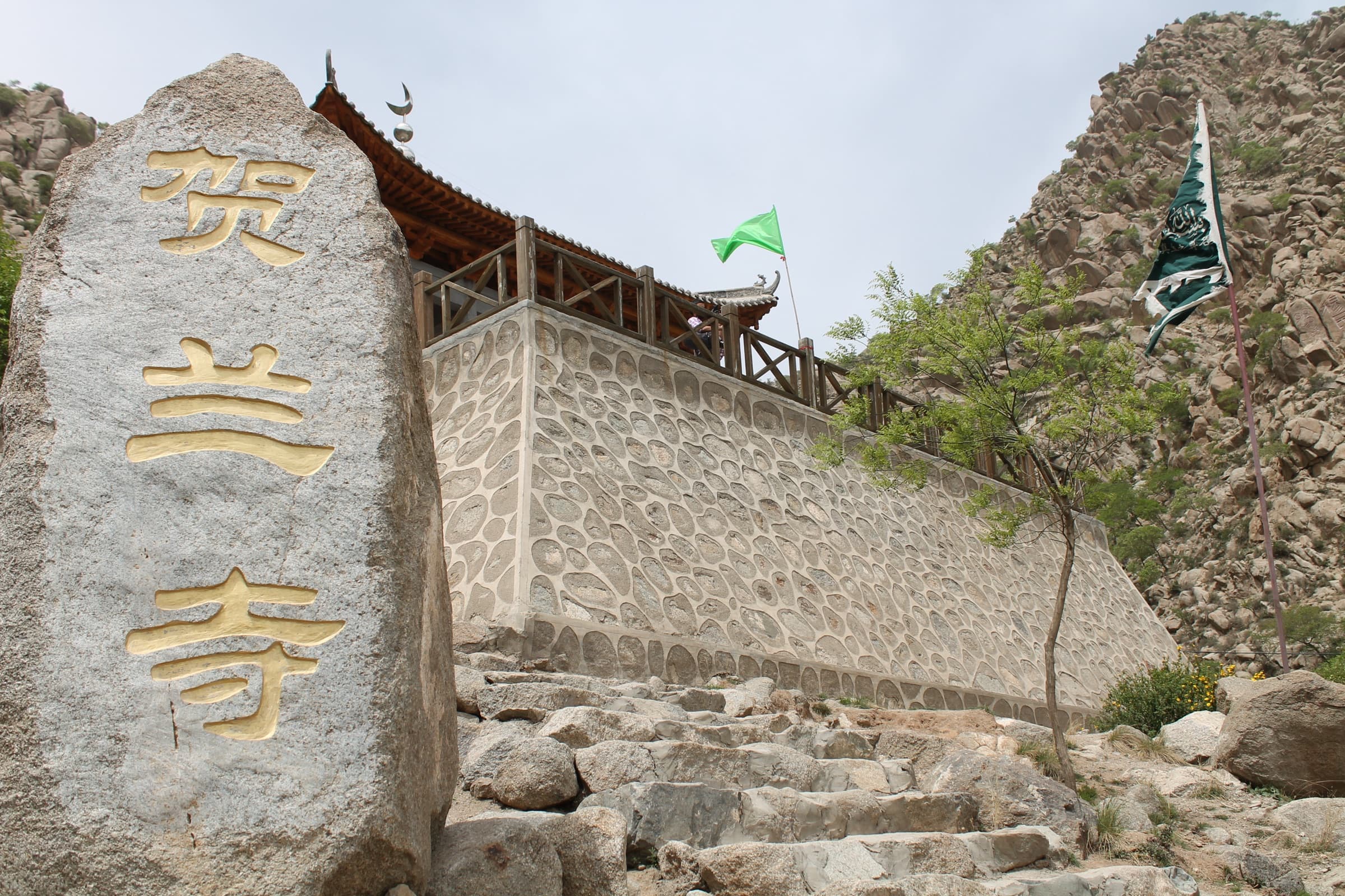 Stèle de pierre gravée de caractères dorés devant un long mur de galets que surmontent des drapeaux verts, adossé à la montagne.