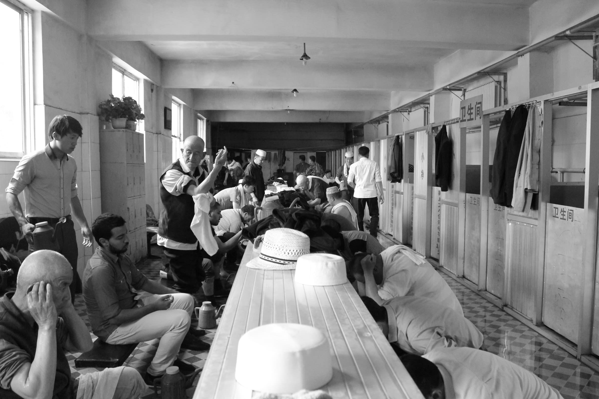 Longue galerie vitrée où des dizaines d’hommes en calot blanc sont prosternés en rangs serrés sur des tapis.