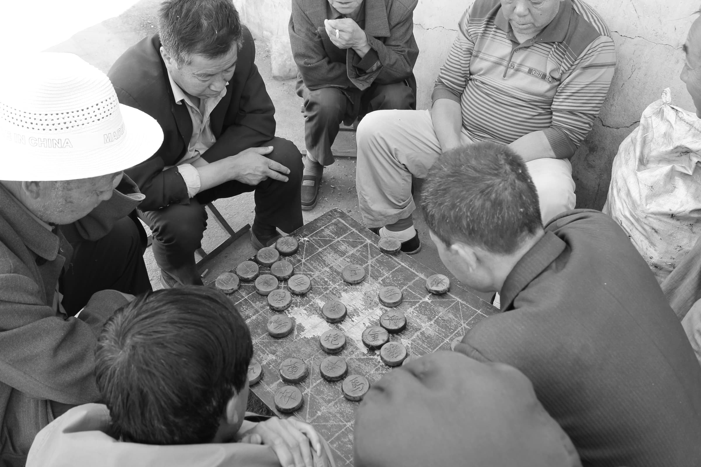 Petit attroupement d’hommes penchés sur un damier posé à même une table basse, dans la rue ; l’un d’eux porte un chapeau de paille clair.