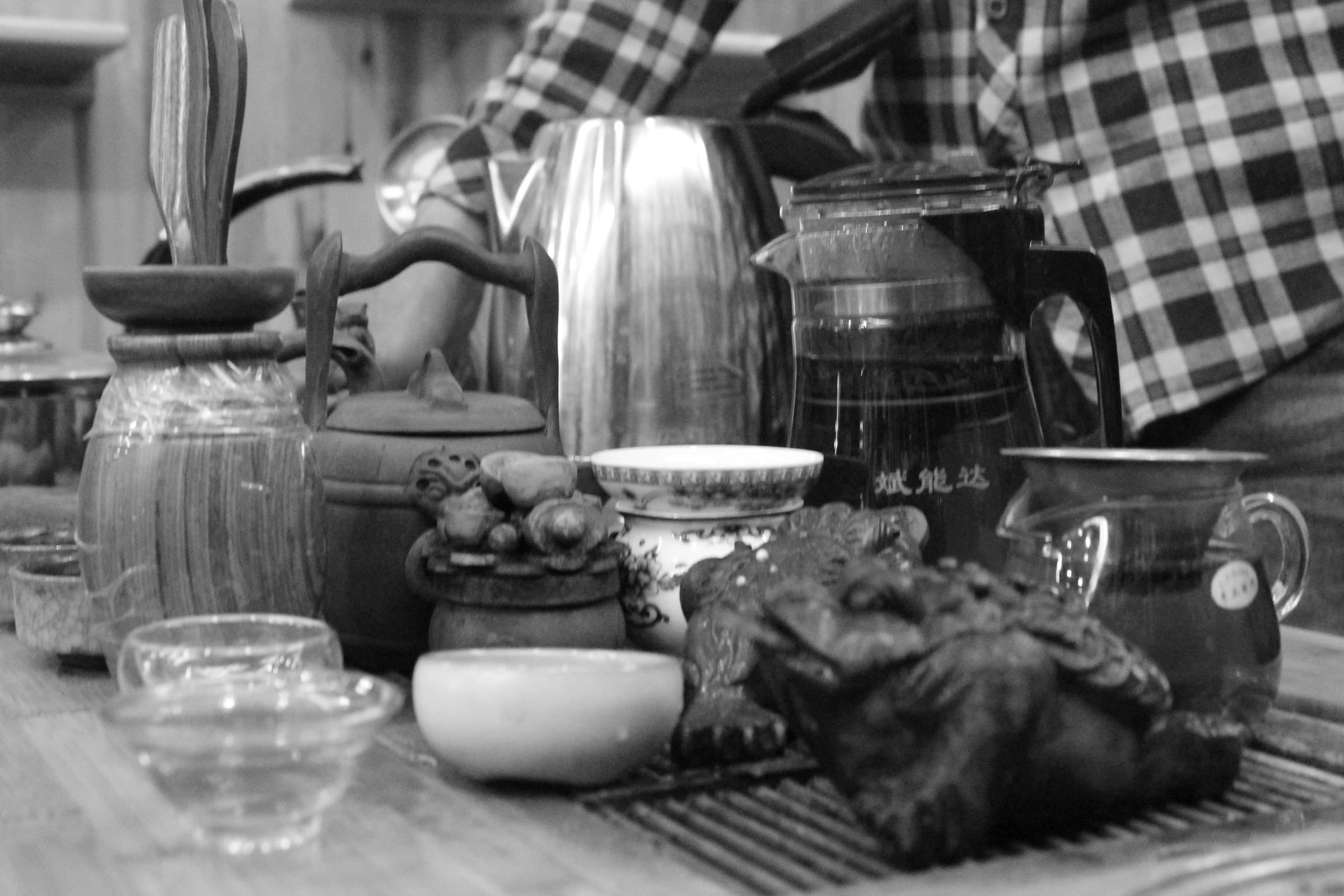 Sur un plateau de bois, une théière de métal, des bocaux de verre et de petites tasses entourent un amas de feuilles ; une main en chemise à carreaux verse l’eau.