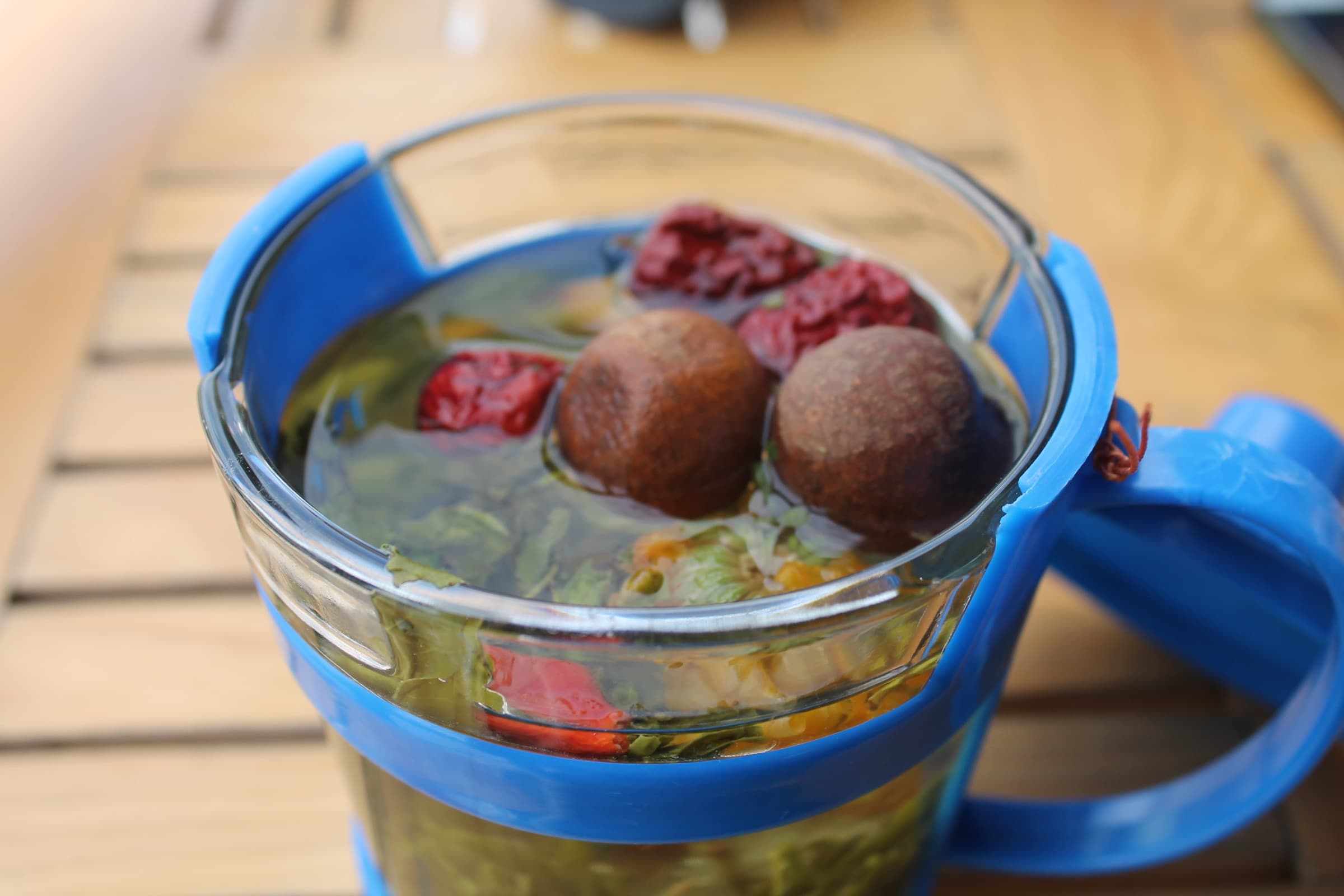 Tasse de verre à monture bleue, remplie d’une infusion où flottent des dattes, des baies rouges et des feuilles de thé vert.