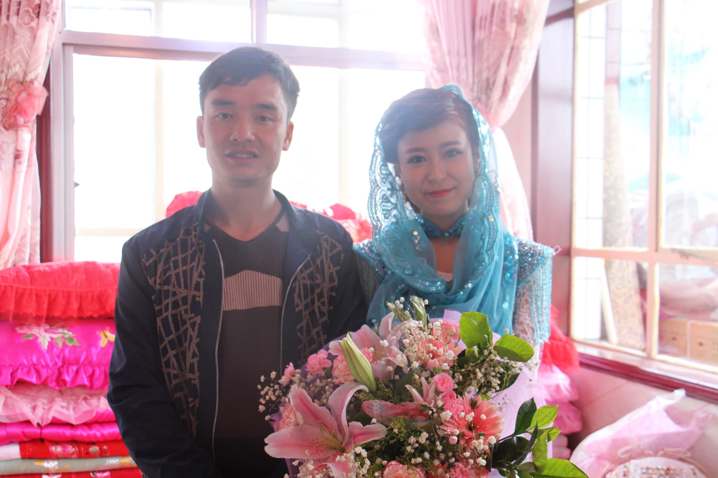 Un jeune couple pose côte à côte, bouquets à la main, devant une tenture et des fleurs de papier rose ; la femme porte un voile de tulle vert.