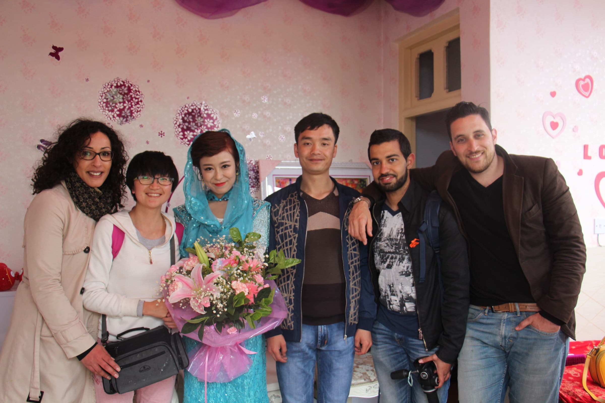 Six personnes posent serrées devant un mur décoré de fleurs roses : les membres de l’équipe entourent la famille et la jeune femme qui tient un bouquet.