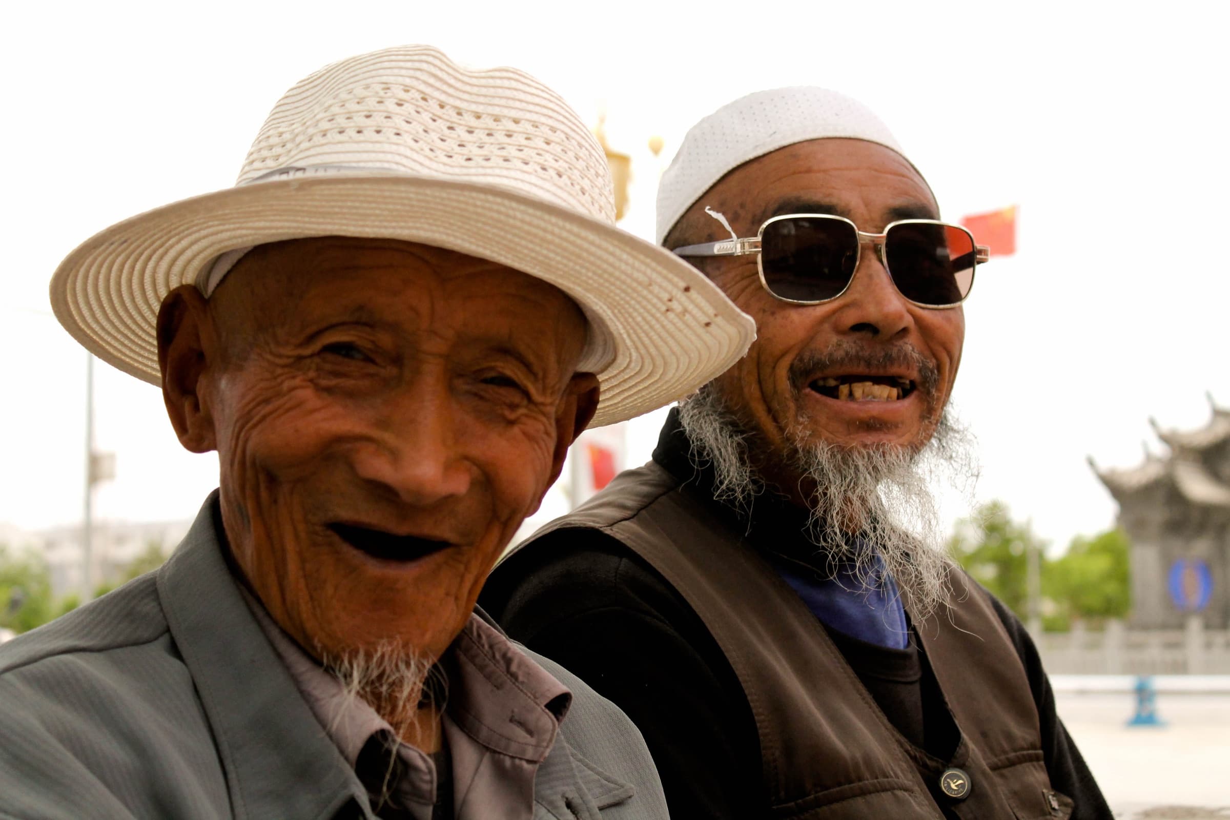 Deux hommes âgés côte à côte, l’un en chapeau de paille clair, l’autre en calot blanc, lunettes noires et longue barbe blanche, riant à pleines dents.