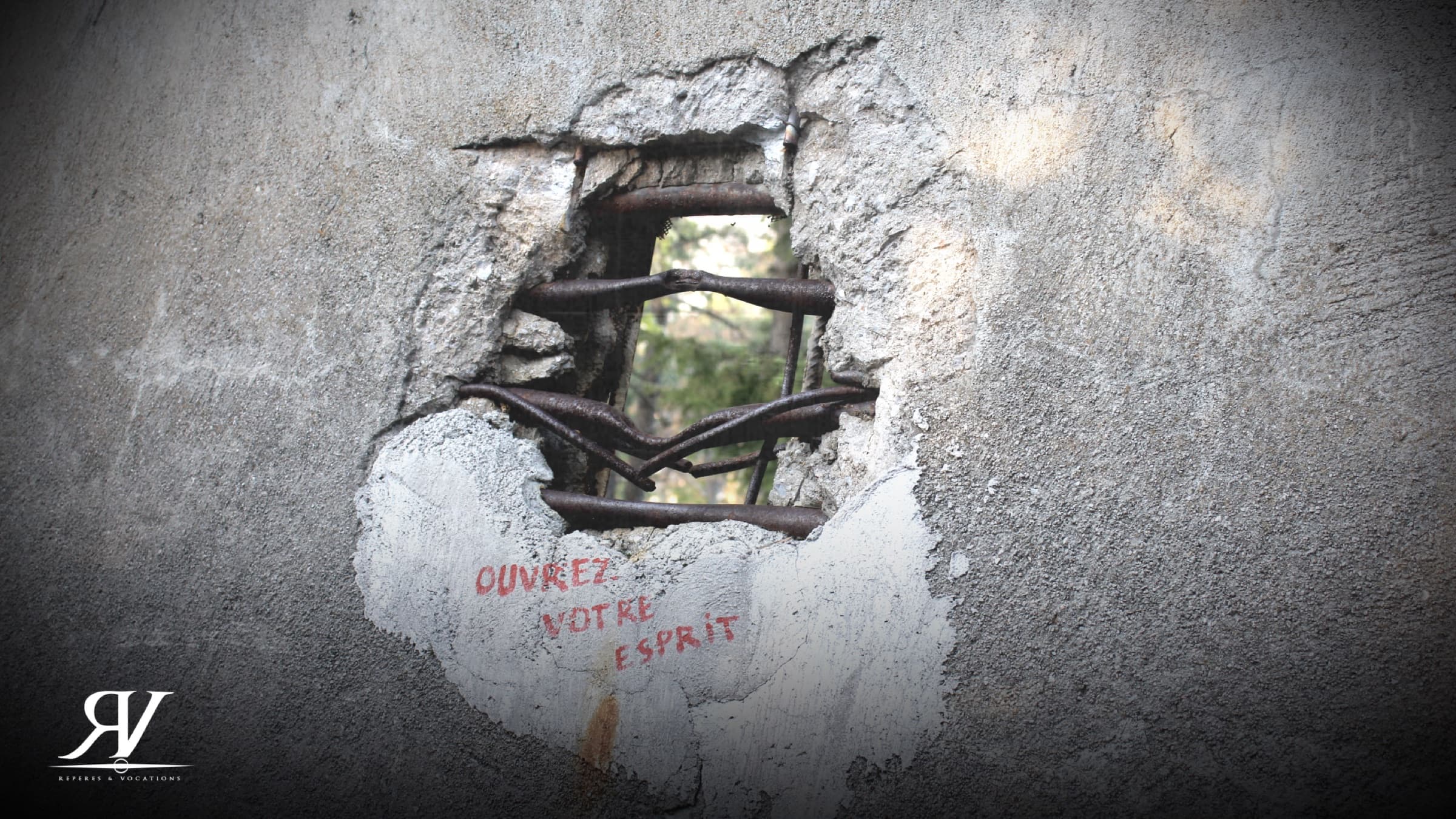 Trou d’obus dans un mur de béton, les fers à béton tordus vers l’extérieur ; en dessous, trois mots peints en rouge : « Ouvrez votre esprit ».