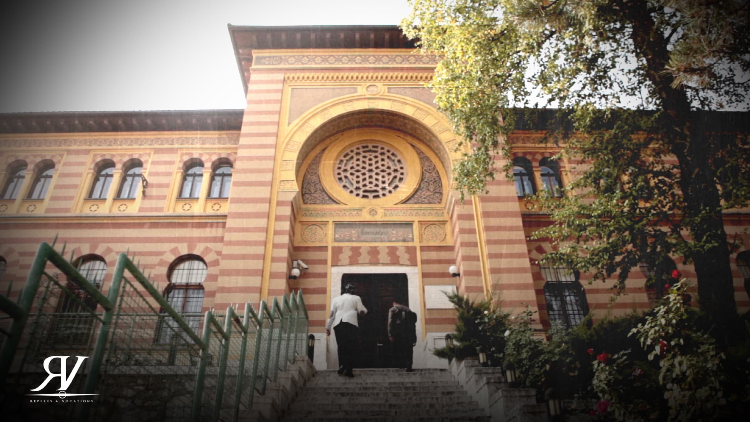 Façade en bandes de pierre ocre et rose d’un bâtiment de style ottoman revisité, grand portail en arc surmonté d’une rosace ; deux personnes gravissent l’escalier d’entrée.