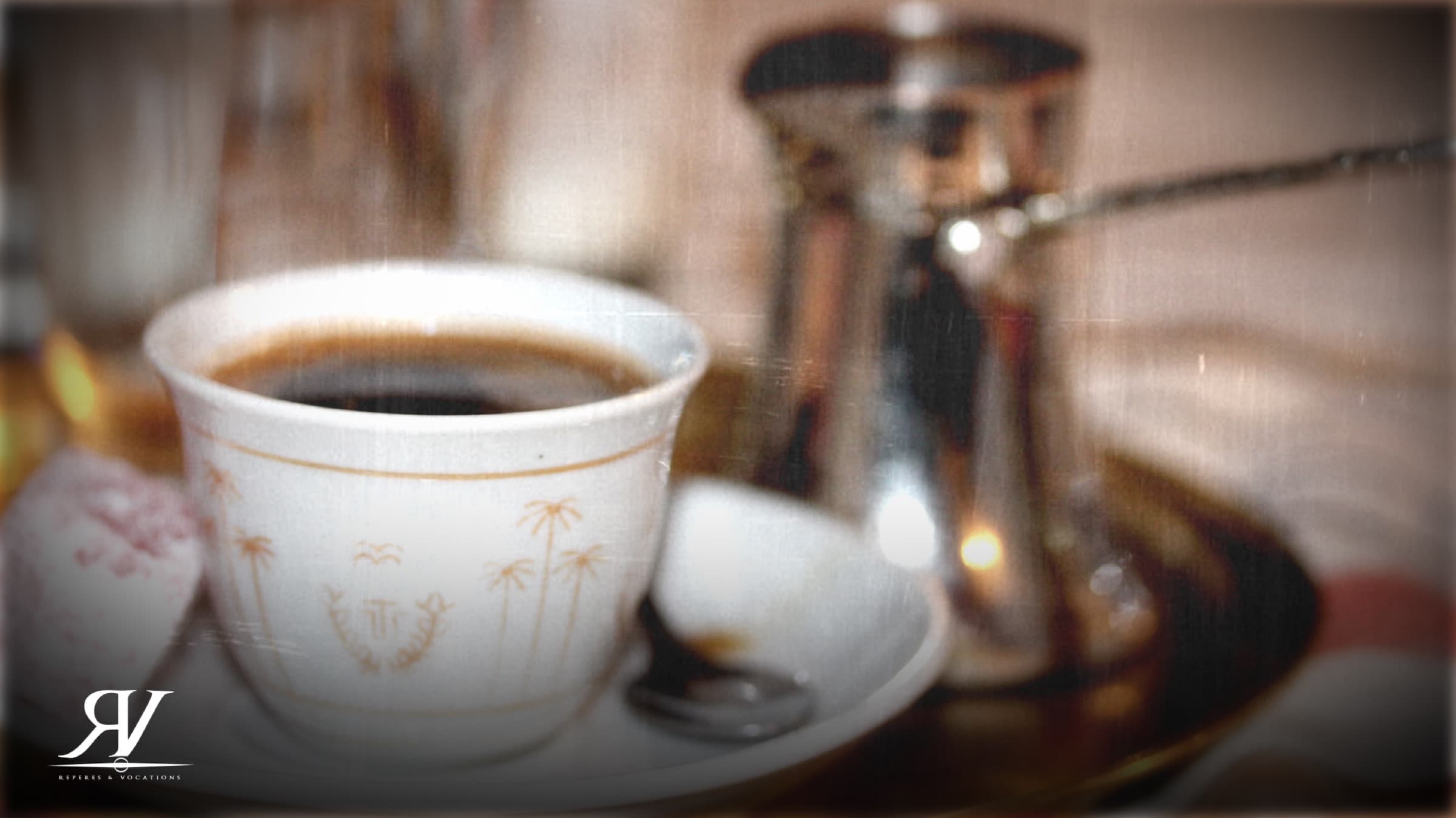 Gros plan sur une tasse de café blanche à motifs dorés posée sur sa soucoupe, une cezve en cuivre brillante derrière elle.