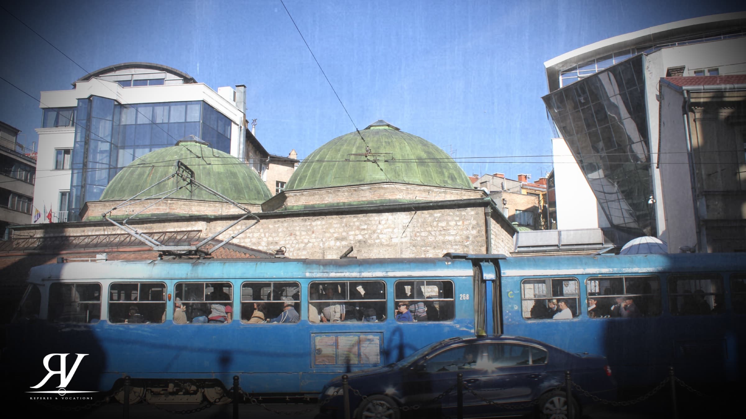 Un tramway bleu passe devant un édifice de pierre couvert de coupoles vert-de-gris, encadré par deux immeubles contemporains en verre.