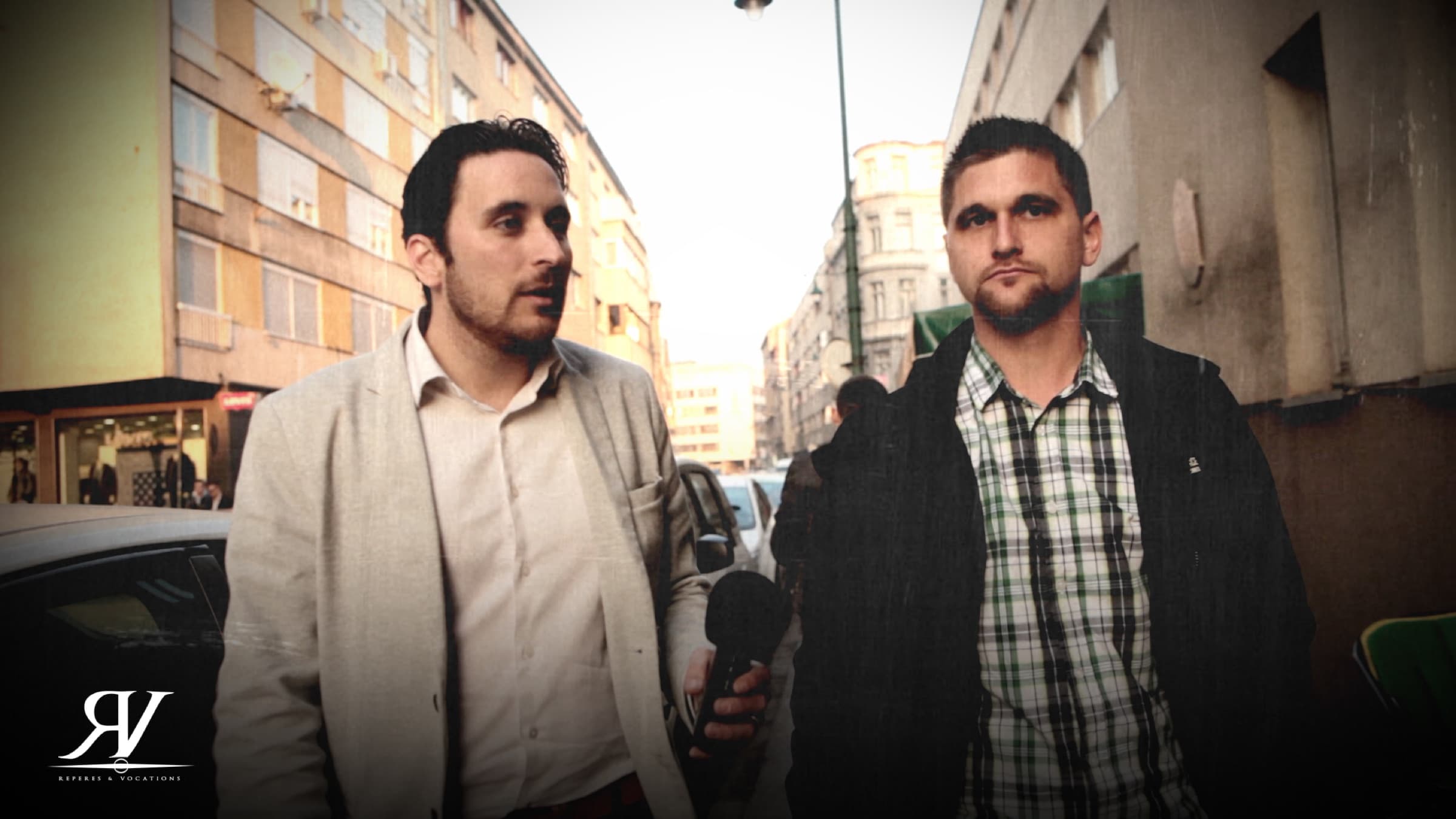 Deux hommes marchent côte à côte dans une rue commerçante ; celui de gauche, en veste claire, tient un micro-main baissé le long du corps.