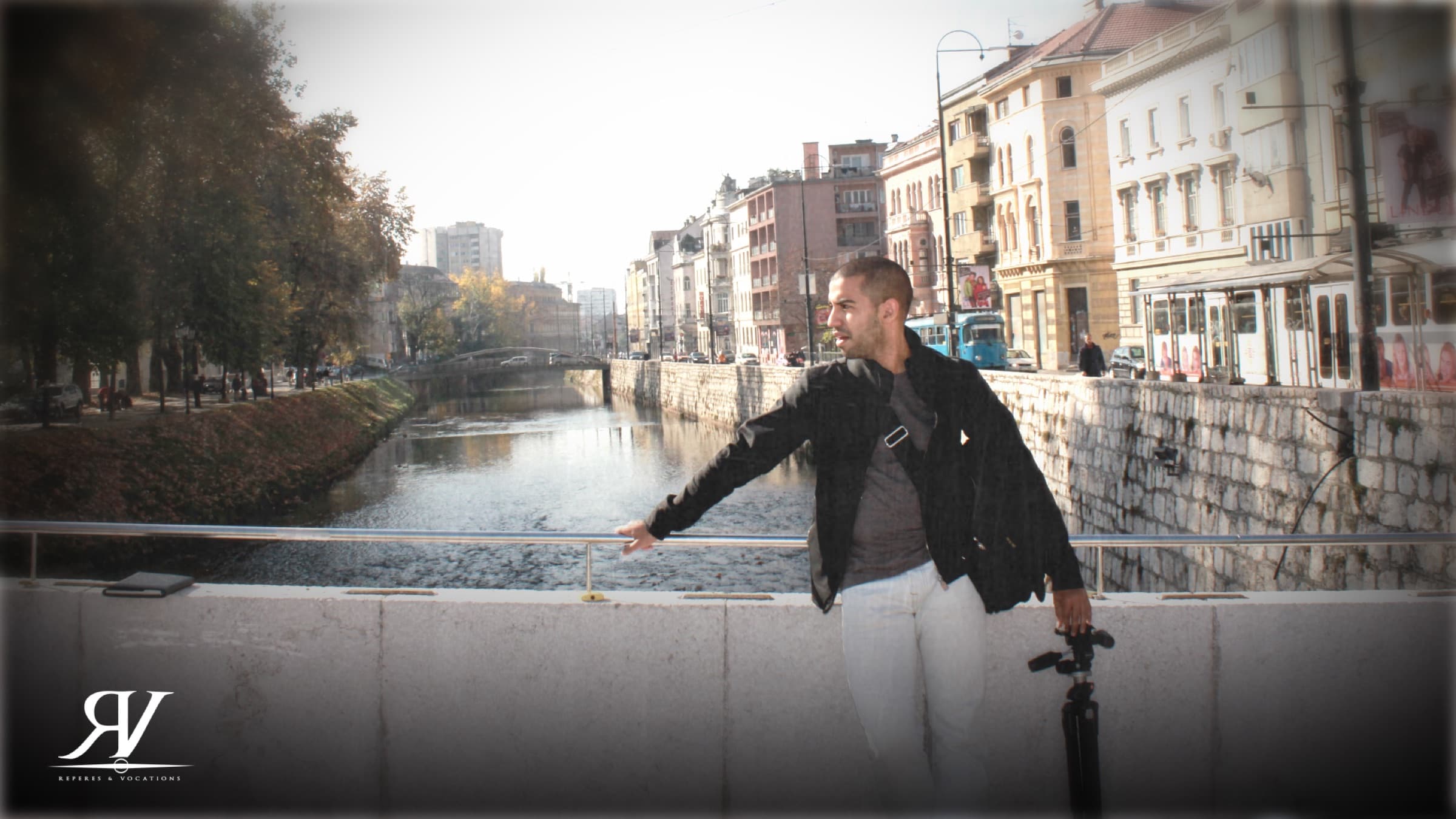 Un homme en veste noire, un pied de caméra à la main, appuyé au parapet d’un quai ; derrière lui, la rivière encaissée et les façades austro-hongroises de Sarajevo.