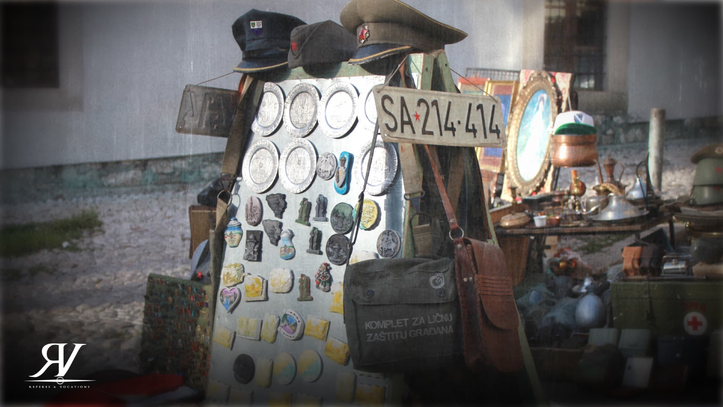 Étal de brocante en plein air : panneau couvert de médailles et d’insignes militaires, casquettes d’uniforme, sacoche kaki et vieille plaque d’immatriculation de Sarajevo.